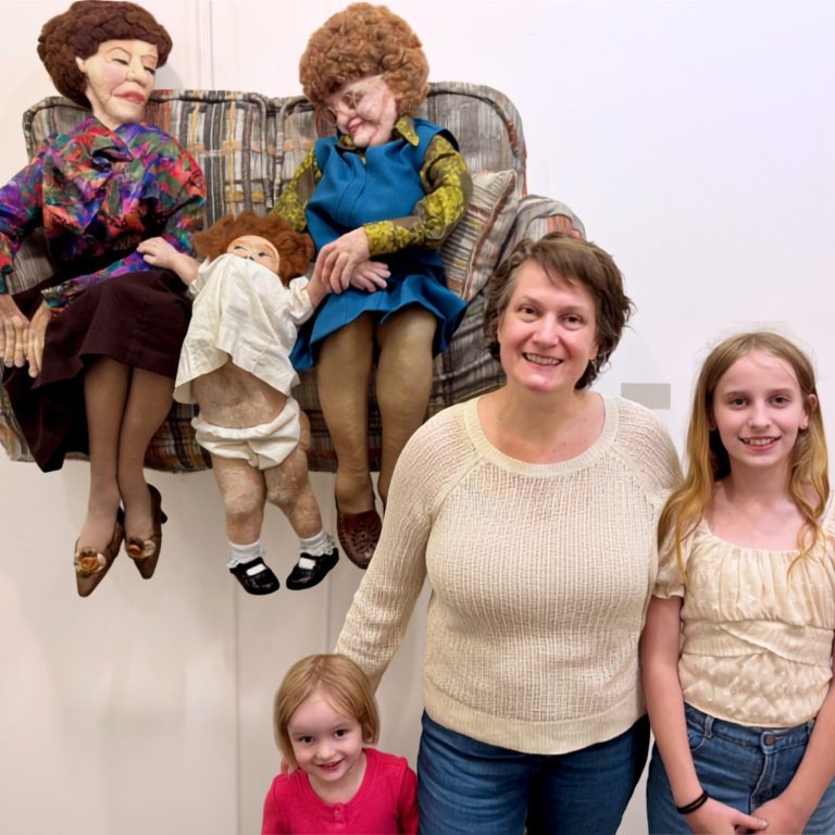 Flint with her sculpture, Inheritance. Artist Emily Flint with her mixed media needle-felted sculpture, Inheritance, depicting two old women and a toddler on a loveseat.