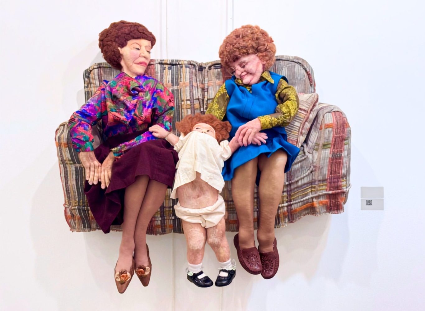 Inheritance, by Emily Flint A mixed-media needle-felted soft-sculpture of two elderly women witha toddler girl squirming free, while sitting on a plaid loveseat.